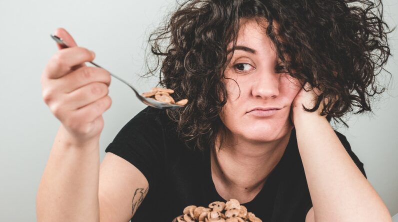 woman with messy hair wearing black crew-neck t-shirt holding spoon with cereals on top